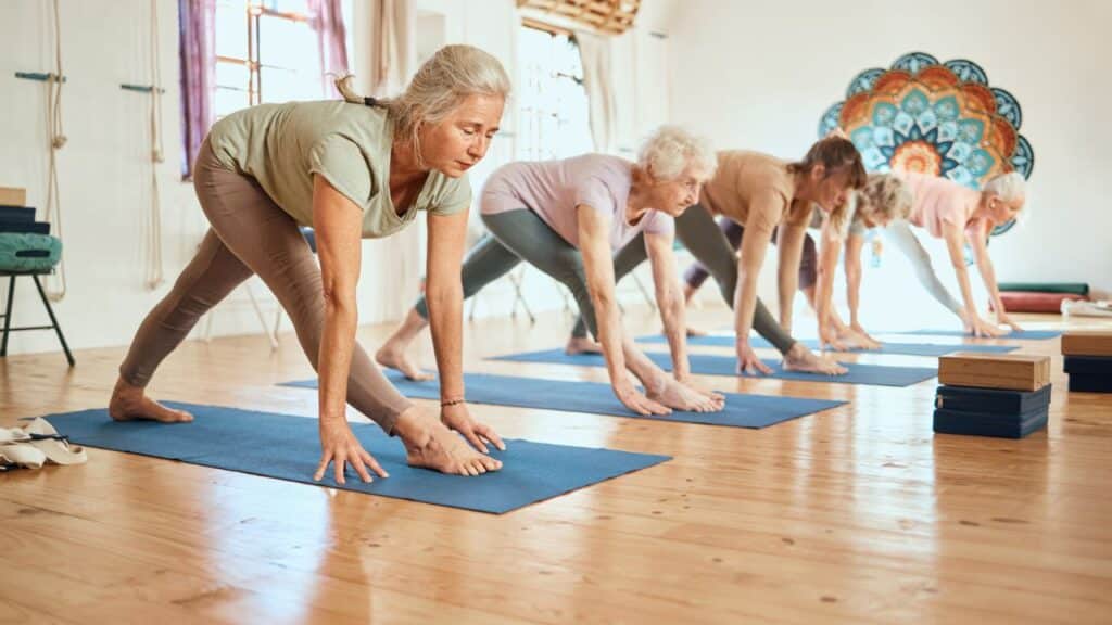 A group of older women in a yoga class bends forward with hands on the floor, stretching on blue mats in a bright studio.