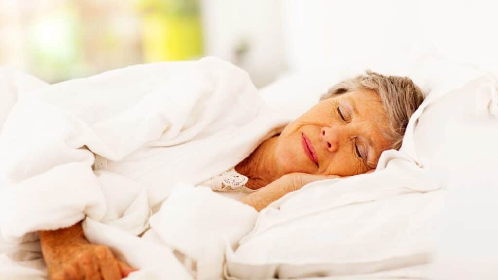 An older woman lies in bed under a white blanket, smiling peacefully with her eyes closed.
