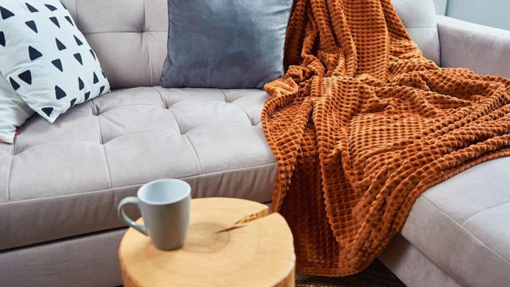 A tufted beige couch with patterned pillows and a soft, textured burnt-orange throw blanket next to a small wood stump coffee table with a gray mug. Ideal lounging for a cozy autumn retreat.