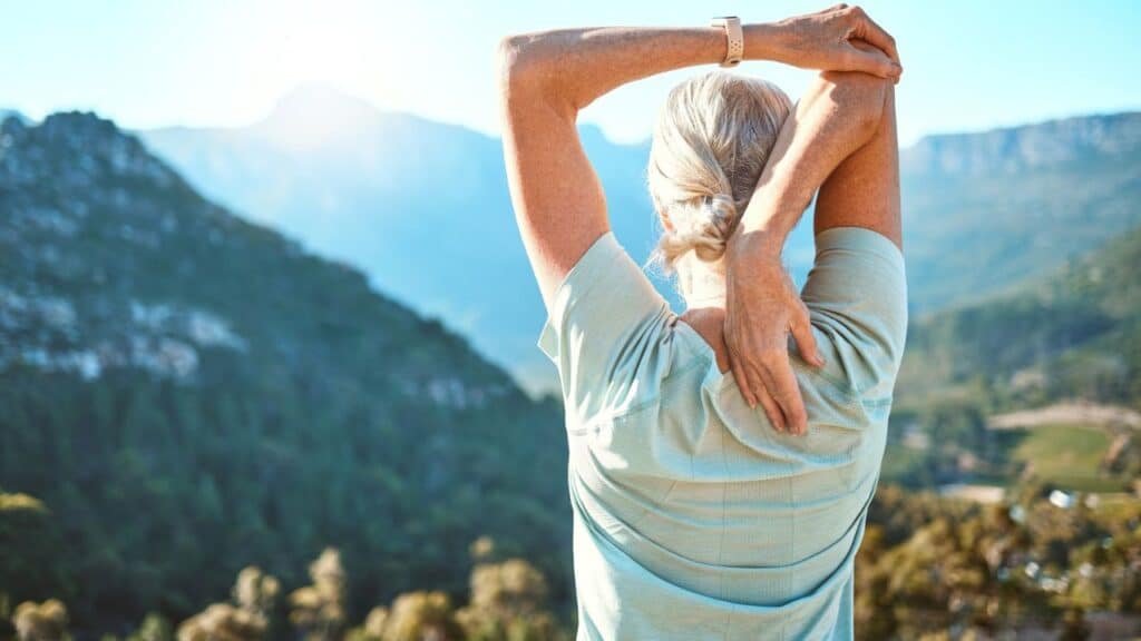 A woman with gray hair stretches her arms overhead while facing a scenic mountain landscape under the sun.