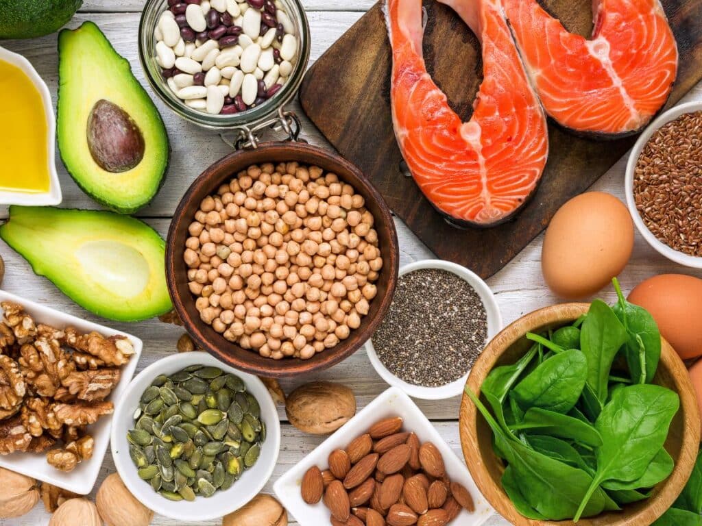 Top view of nutrient-rich foods including salmon, chickpeas, avocado, eggs, spinach, seeds, and nuts arranged on a table. These whole foods are packed with antioxidants and omega-3s, making them essential components of anti-aging secrets for skin and body health.