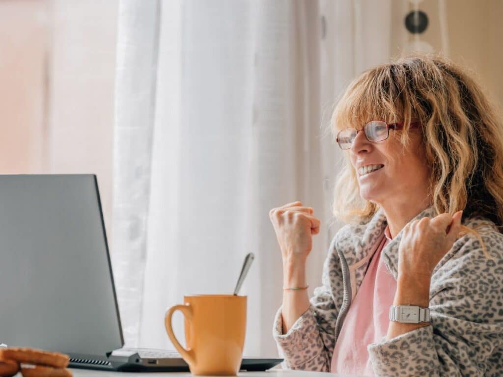 A smiling woman in pajamas pumps her fists in celebration while looking at her laptop, sitting at a breakfast table with a mug. Reflects the excitement of success from smart side hustles at home.