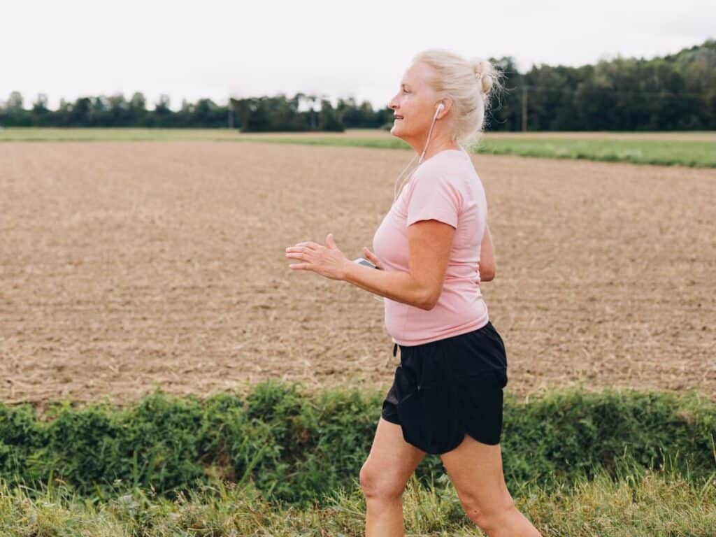 Older woman in athletic wear jogging along a rural path with earphones in, smiling as she exercises. Regular physical activity like this is one of the most effective anti-aging secrets for maintaining vitality and overall wellness.