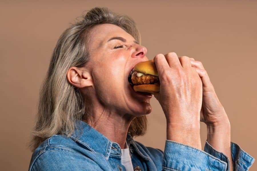 A mature woman wearing denim takes a large bite of a burger against a neutral background. Eating heavy meals late at night can interfere with nightly rituals that rewind the clock on aging.