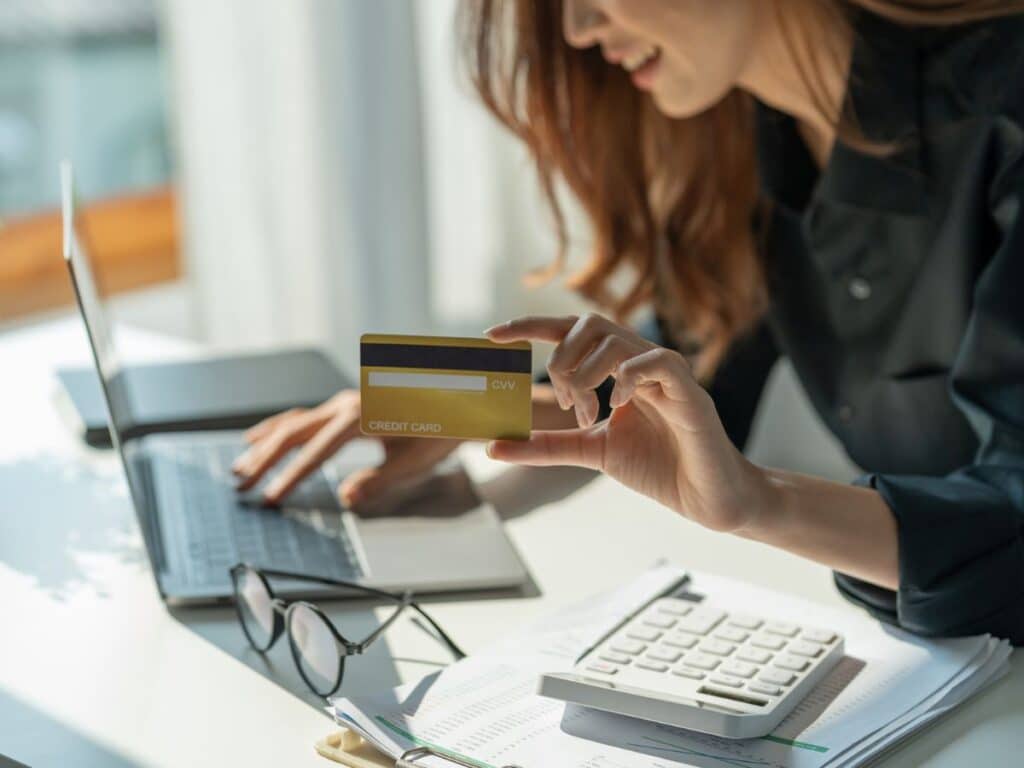 A woman smiles while holding a gold credit card in one hand and typing on a laptop with the other, with documents, glasses, and a calculator on the table.