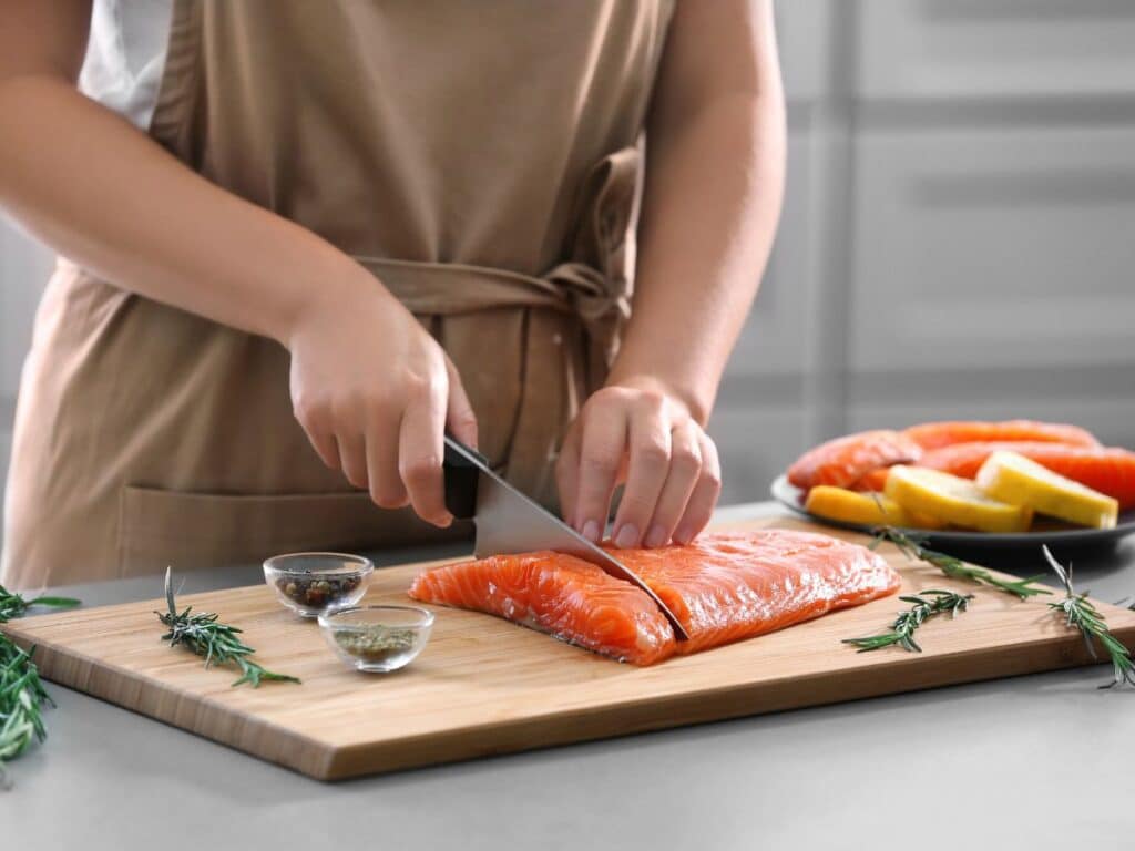 A person wearing an apron slicing raw salmon on a wooden board with small bowls of seasoning and herbs nearby. Preparing serotonin-boosting salmon for a healthy meal.
