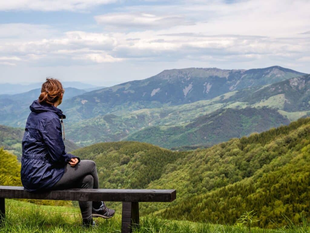 Woman in outdoor gear sitting on a bench and admiring a lush green mountain landscape. Regular exposure to nature and stress-reducing habits like this are among the often-overlooked anti-aging secrets for mental and physical well-being.