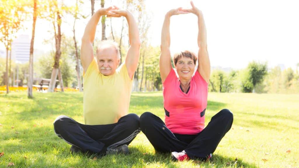 An older man and woman sit on green grass in a sunny park, both stretching their arms straight above their heads.