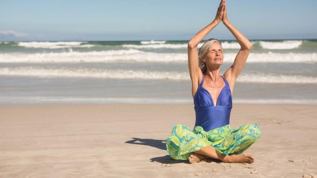 An older woman in a blue swimsuit top and green patterned pants sits cross-legged on the beach with her hands pressed together above her head.