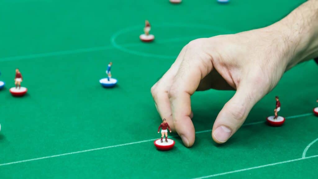 A hand positions a red-uniformed Subbuteo soccer figure on a green tabletop pitch, surrounded by other tiny players. This flick-to-kick game was a standout among childhood toys from the 70s for sports lovers.