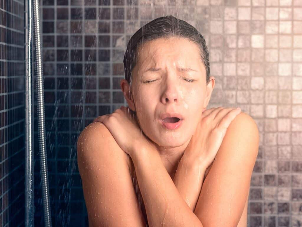 Woman reacting to a cold shower with arms crossed over her chest and eyes closed under running water. Cold water therapy is gaining popularity as one of the bold anti-aging secrets for improving circulation and skin tone.