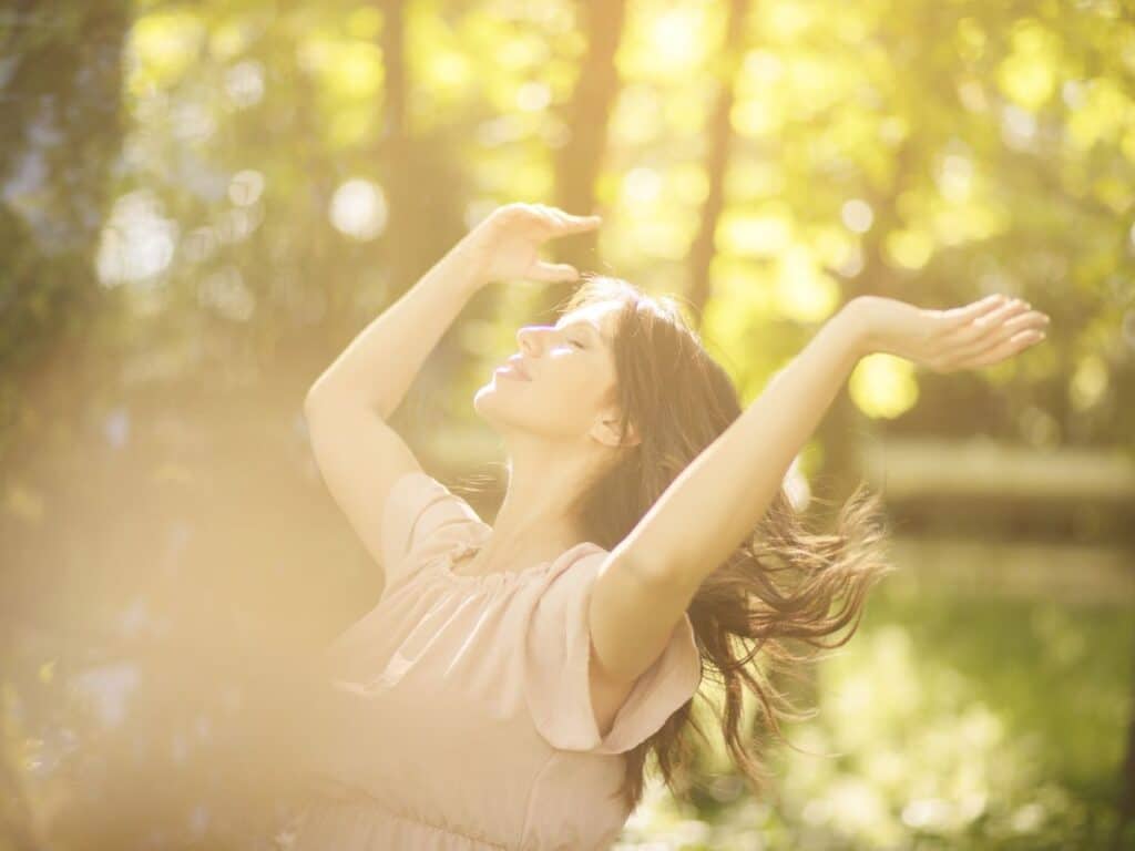 Woman with long hair basking in warm sunlight with arms raised and eyes closed in a forest. Controlled sun exposure helps vitamin D production and is considered one of the natural anti-aging secrets when done safely.