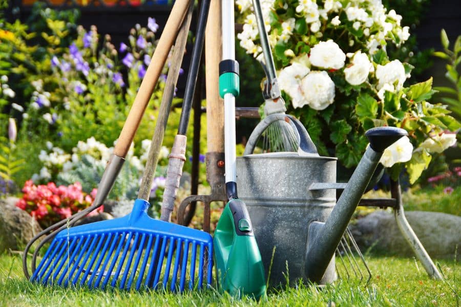 A group of garden tools including rakes, a watering can, a hoe, and pruning shears stands upright in lush grass near blooming flowers, positioned like a ready-to-go Facebook Marketplace bundle.