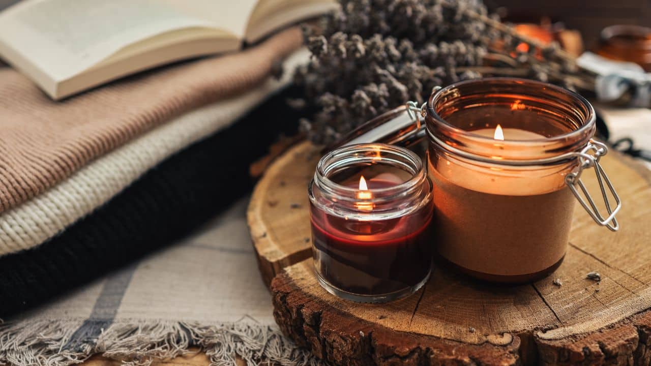 Two lit candles in glass jars rest on a rustic wood slice, surrounded by dried lavender and folded knit blankets with an open book in the background. A warm and relaxing setup perfect for a cozy autumn retreat moment.
