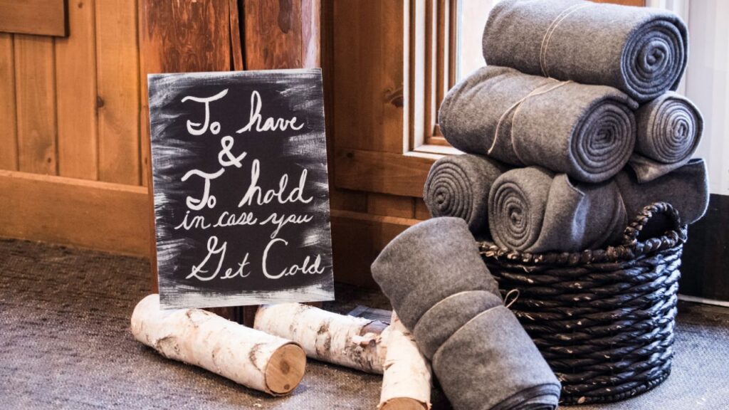 Rolled gray wool blankets stacked in a woven basket beside a chalkboard sign that reads “To have & To hold in case you get cold,” with birch logs on the floor. Inviting touch for guests at a cozy autumn retreat or rustic event.