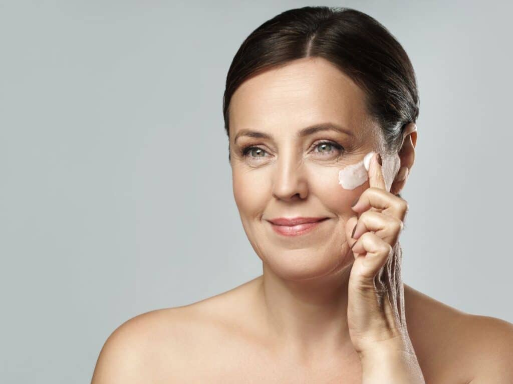 Mature woman with bare shoulders gently applying face cream to her cheek while smiling. Consistent moisturizing is a foundational step in many anti-aging secrets to maintain elasticity and glow.