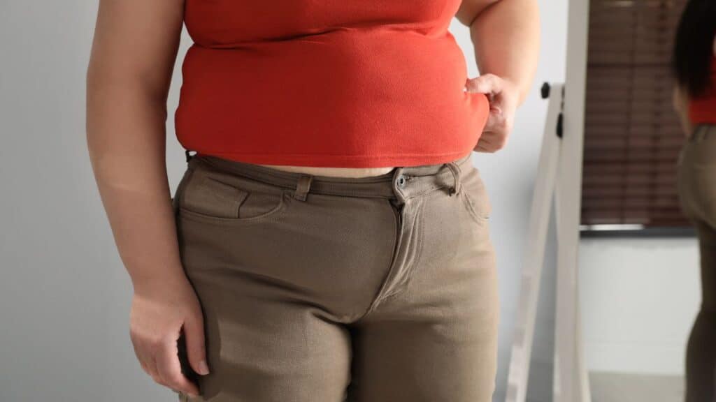 Close-up of a person struggling to button tight brown pants with a red top, highlighting an uncomfortable fit that is a classic fashion faux pas.