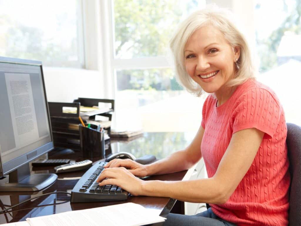 A cheerful woman in a coral sweater types on a desktop computer in a bright home office. Represents engaging in smart side hustles like freelance writing or virtual assistance.