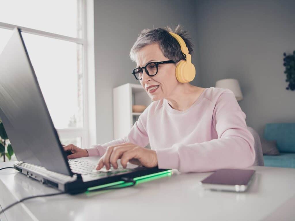A focused woman with short gray hair and yellow headphones works intensely on a gaming-style laptop. Captures tech-savvy smart side hustles like streaming or editing