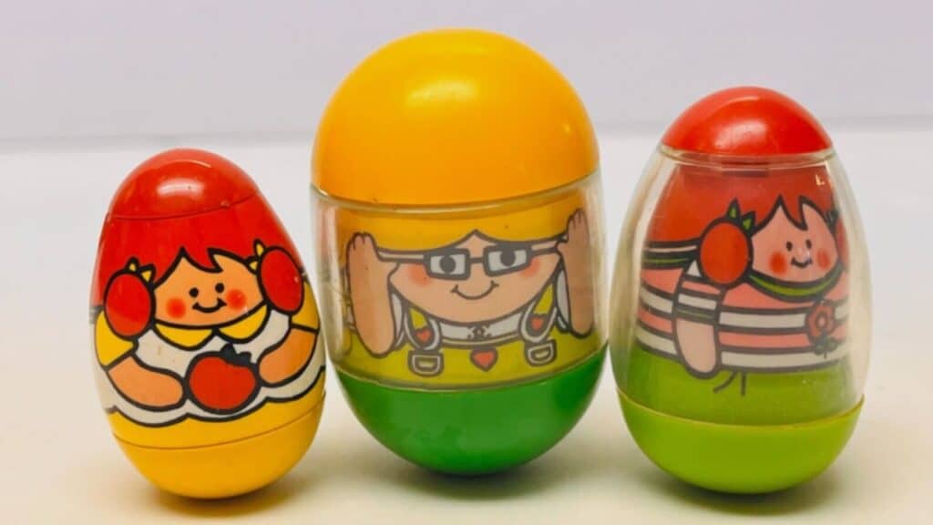 Three colorful egg-shaped Weebles with smiling cartoon faces stand upright, featuring red, yellow, green, and orange accents. Known for never falling down, these wobbling characters were iconic childhood toys from the 70s.