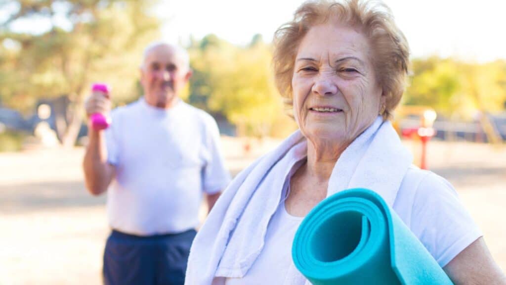 An older woman smiles while holding a rolled-up blue yoga mat and wearing a white towel around her neck, with an older man in the background holding pink dumbbells.