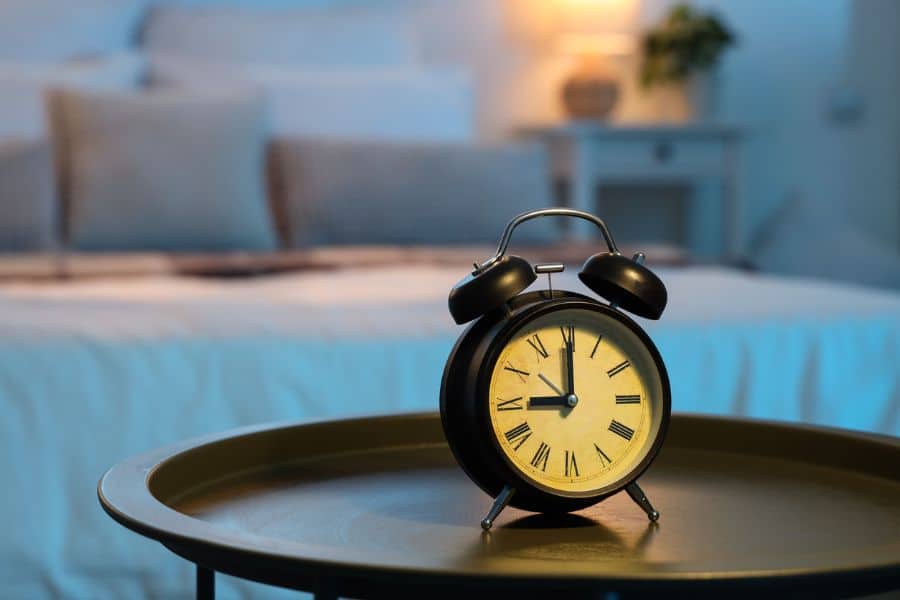 A vintage black alarm clock showing 10:10 on a bedside table with a softly lit bedroom in the background. Keeping a consistent bedtime routine supports nightly rituals that rewind the clock on aging.