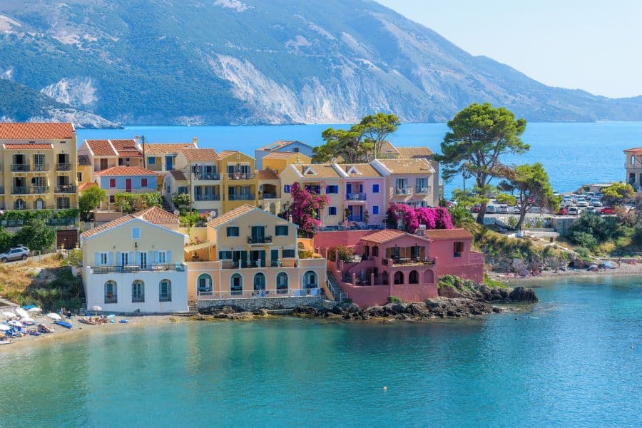 Colorful coastal homes in Assos, a village on the Greek island of Kefalonia, clustered along turquoise waters with dramatic mountains in the background. A prime example of European villages on the coast blending architecture and nature.