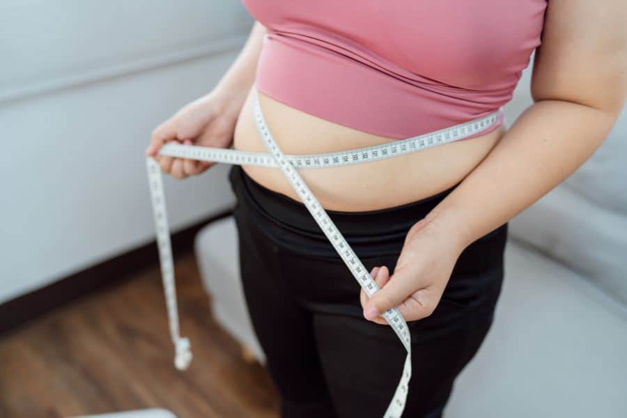 Woman in a pink top with her belly showing and black pants with a measuring tape around her stomach area.