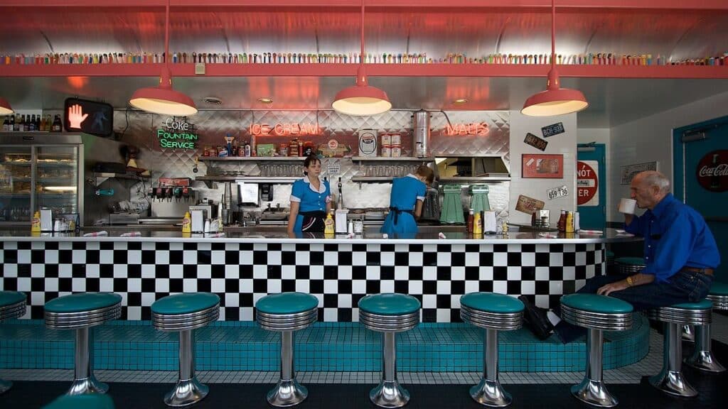 Retro-style diner interior with black-and-white checkered counter, teal barstools, and two waitresses in blue uniforms, serving a customer at the counter. A classic example of nostalgic restaurants on Route 66.