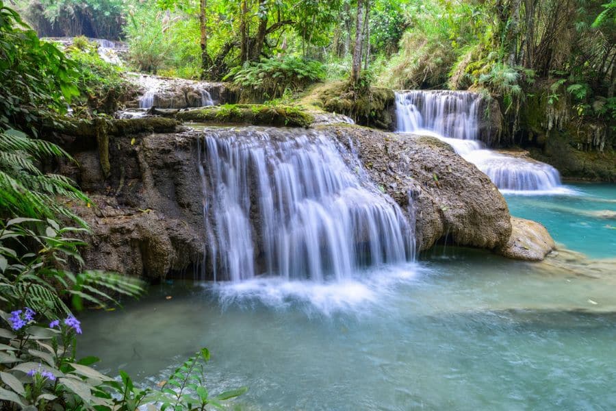 Multi-tiered waterfall cascading over smooth rocks into a turquoise pool, surrounded by dense green vegetation and tropical plants.