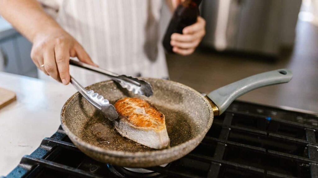 A person uses metal tongs to flip a salmon steak in a frying pan, showcasing essential kitchen tools in action during cooking.