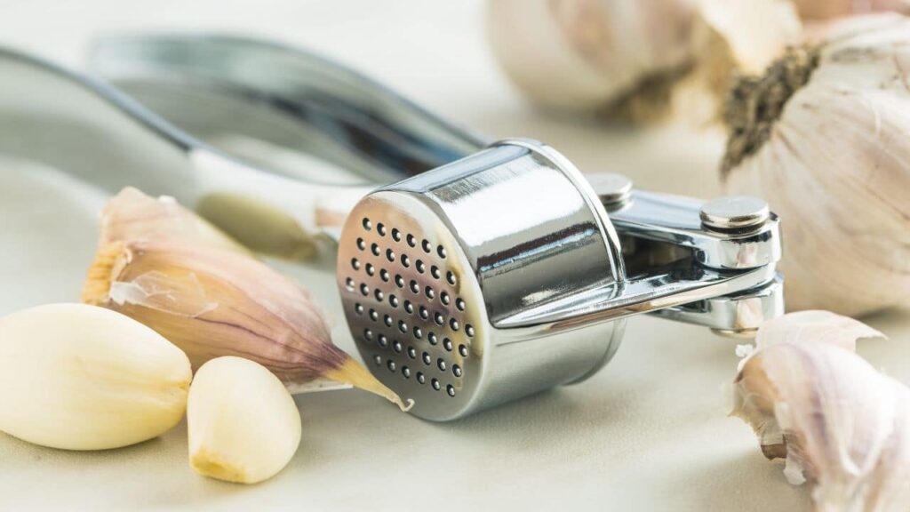 A garlic press surrounded by fresh garlic cloves on a light-colored surface, highlighting one of the smaller yet useful kitchen tools.
