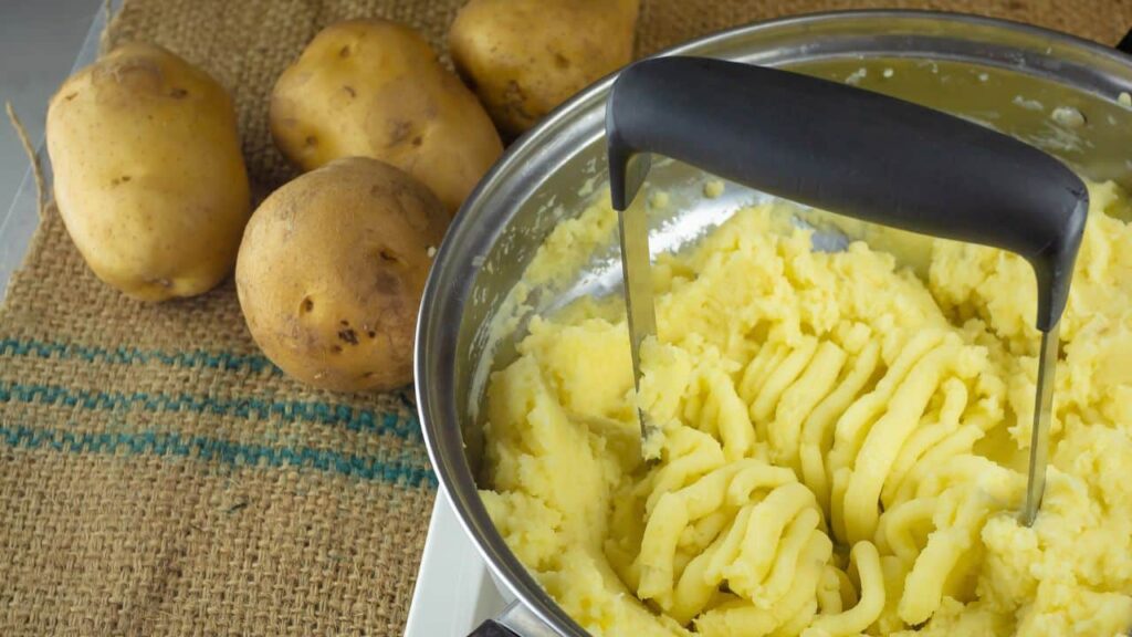 A stainless steel bowl of mashed potatoes being mixed with a potato masher, with whole potatoes nearby, demonstrating manual kitchen tools for food prep.