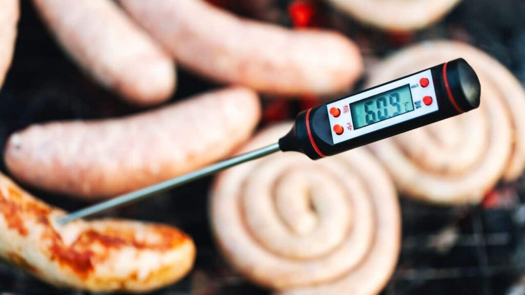 A digital meat thermometer inserted into a sausage on a grill, showing precision kitchen tools for checking internal food temperature.