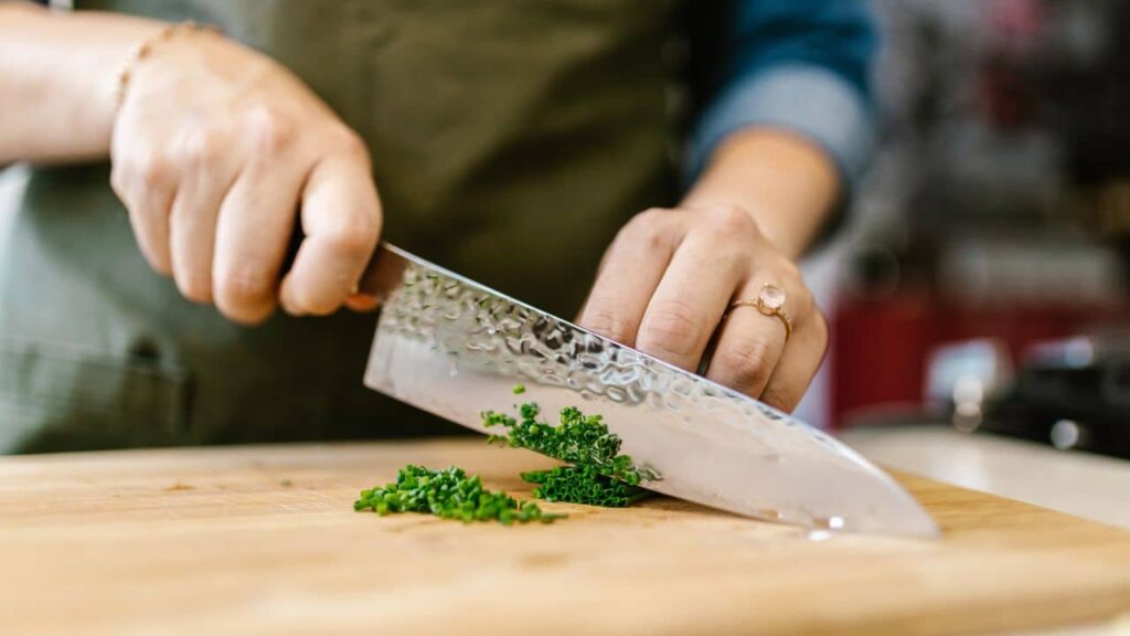 A close-up of hands chopping herbs with a large kitchen knife on a wooden board, showcasing basic kitchen tools for cutting and meal prep.