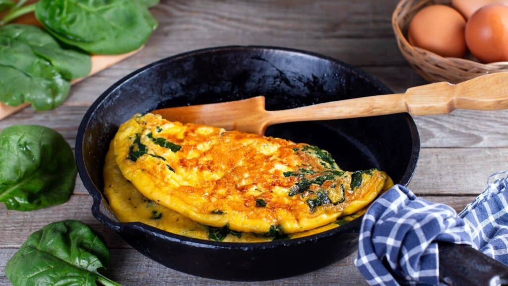 A spinach and cheese omelet cooking in a cast iron skillet with a wooden spatula, surrounded by fresh spinach and eggs, highlighting classic kitchen tools for stovetop cooking.