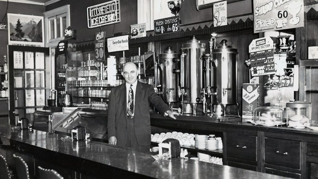Black-and-white photo of a man in a suit behind a vintage soda fountain counter, surrounded by signs advertising chili, oysters, and cocktails. A historic glimpse into early restaurants on Route 66.