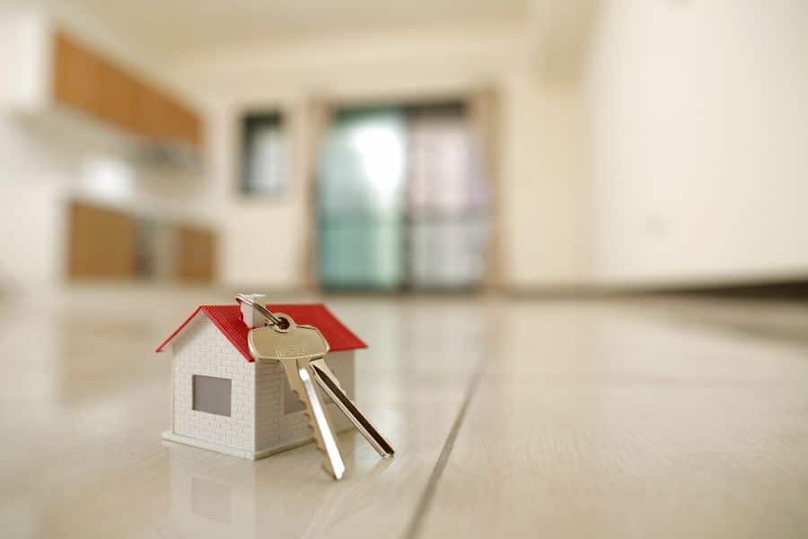 A tiny house model with a red roof and silver keys placed on a clean floor in an empty room. Symbolizes property purchase and key handover in real estate acquisition strategies.