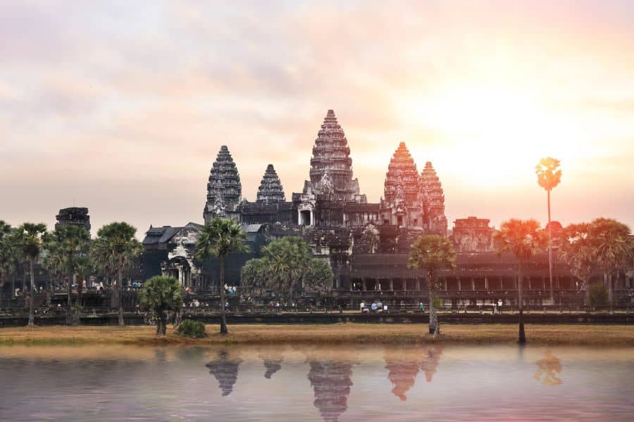 Majestic view of Angkor Wat temple complex silhouetted against a glowing sunrise sky, with palm trees and reflection in a still body of water.