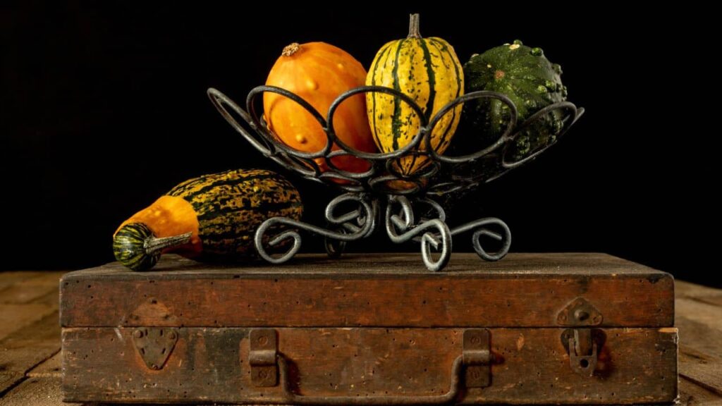 A rustic metal basket holding a variety of colorful gourds, placed on a vintage wooden chest against a dark background. This harvest-themed arrangement complements fall porch decor with natural, earthy elements.