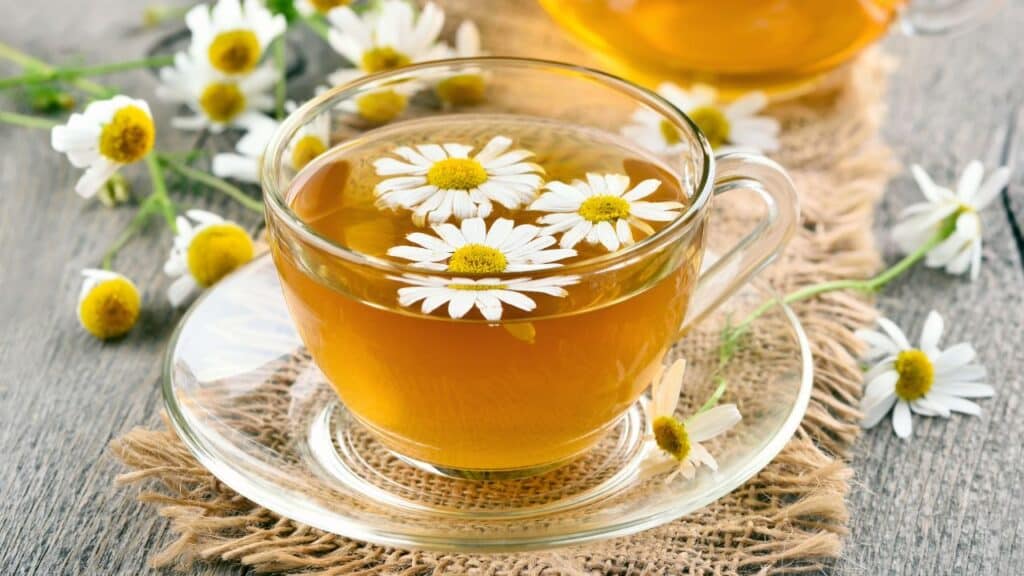 A clear glass cup of chamomile tea with fresh chamomile flowers floating on top, surrounded by loose blooms on a wooden table. A classic herbal drink featured in vintage remedies for calming effects.