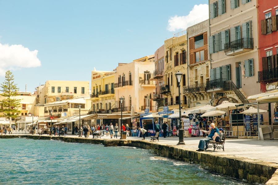 People strolling along the Venetian harbor of Chania in Crete, Greece, with pastel buildings, seaside cafes, and a vibrant, historic atmosphere. A lively scene from one of Europe's most charming coastal villages.