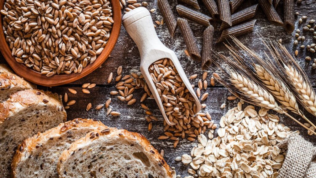 A variety of whole grains including oats, whole grain bread, wheat stalks, and dark pasta displayed on a wooden table. A great visual for diet tips emphasizing fiber and heart-healthy carbs.