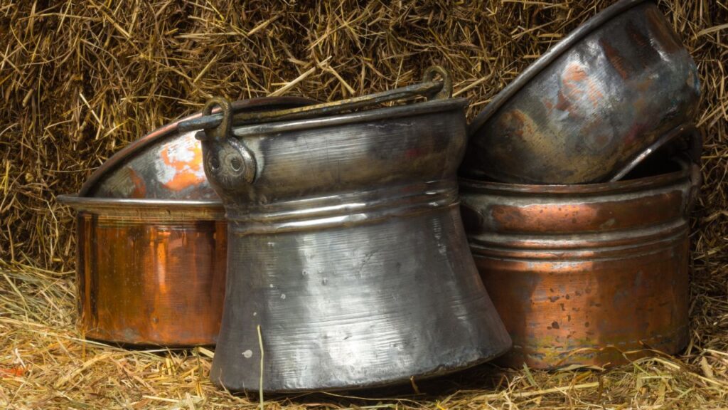 A set of aged copper and metal pots leaning against a hay bale with visible scratches and patina. These rustic containers bring a vintage farm vibe to fall porch decor.