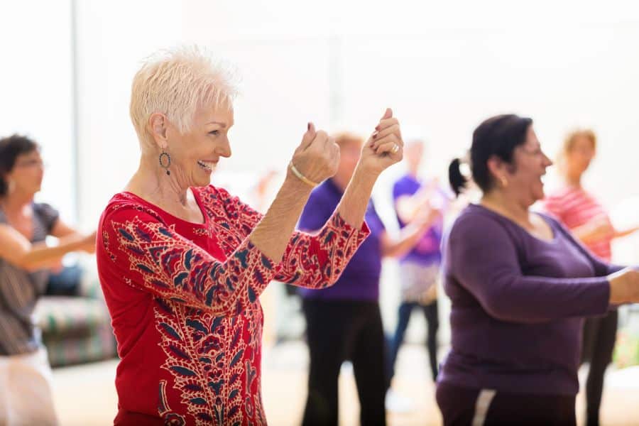 Senior woman smiling and dancing in a group fitness class, wearing a red patterned top. Dance classes offer fun and social everyday exercises for older adults.