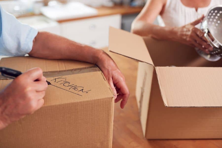 Two people packing kitchenware into cardboard boxes, with one box labeled “KITCHEN” in marker. Illustrates the moving process often involved in property flipping or relocation strategies.