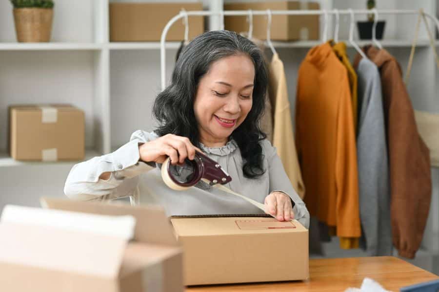 A smiling older woman tapes a cardboard box closed in a home workspace, illustrating small-scale online selling or packaging as a retiree-friendly job option.