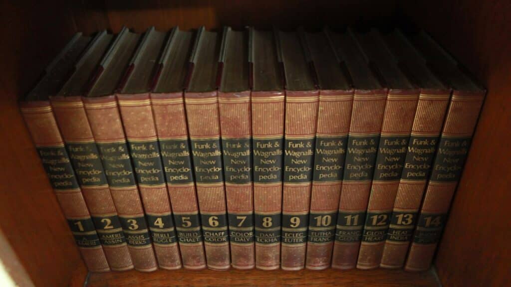 A full shelf of Funk & Wagnalls New Encyclopedia volumes lined up in order with red and gold spines in a wooden bookcase.