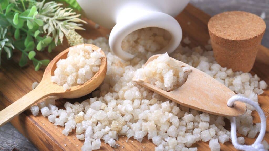 Coarse bath salts in wooden scoops and spilling from a ceramic jar, styled with dried greenery. Bath soaks like this are often part of vintage remedies for relaxation and detoxification.