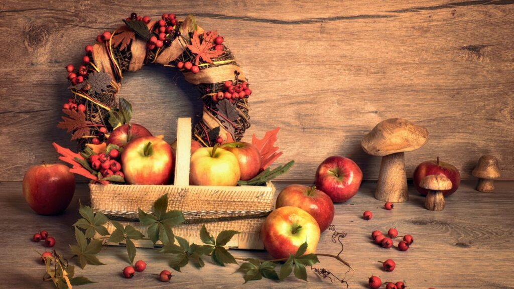 A basket of apples and red berries sits in front of a wreath made of twigs, leaves, and ribbon, alongside decorative mushrooms on a wooden surface. This harvest-style arrangement suits warm and organic fall porch decor.